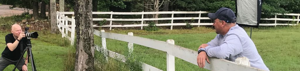 A photographer takes a portrait of a man leaning on a fence.